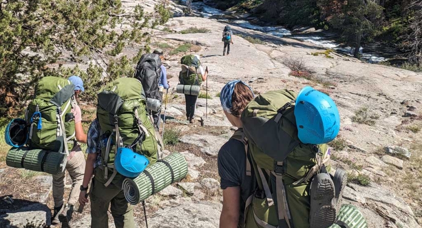 A group of people wearing backpacks hike away from the camera over a rocky landscape. 